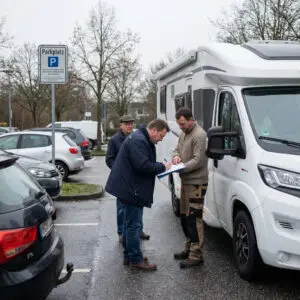 Zwei Männer unterzeichnen auf einem Parkplatz informell ein Dokument an der Seitentür eines weißen Wohnmobils.